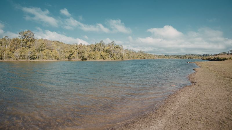 Wide sandy shoreline along a calm lake lined with autumn-toned trees under a partly cloudy sky.