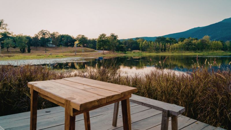 Wooden table and bench on a deck overlooking a calm lake with tall grass, trees, and a mountain at dusk.