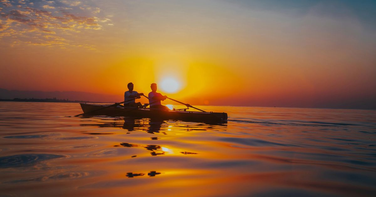 Two paddlers silhouetted in a kayak on calm water at sunset with a vivid orange sky.