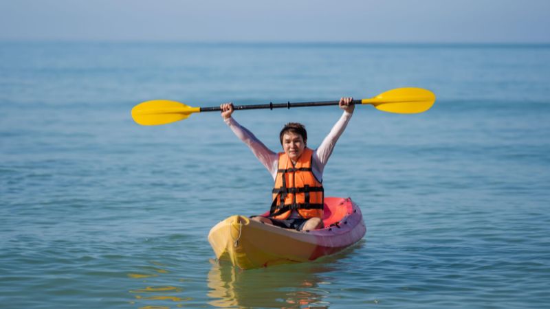 A kayaker in an orange life jacket holds a yellow paddle overhead while seated in a yellow kayak on calm open water.