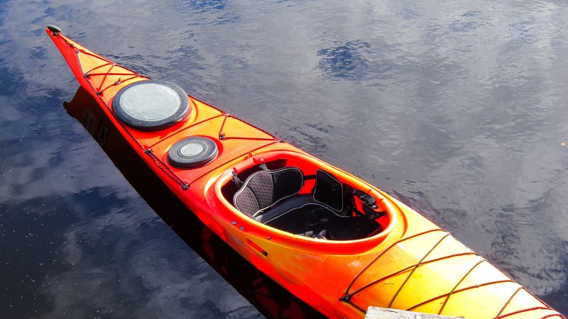 An orange and yellow sea kayak with hatch covers floats on still, dark water.