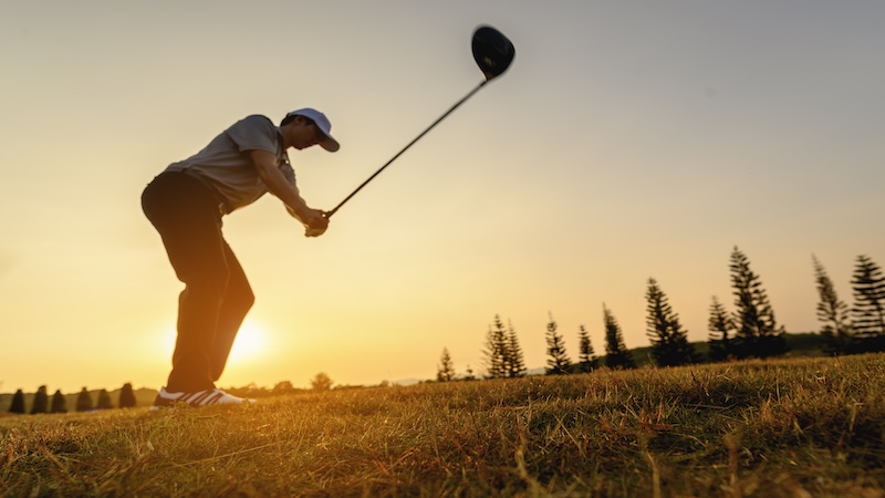 Golfer swinging a club on a golf course at sunset with golden sky and trees in the background