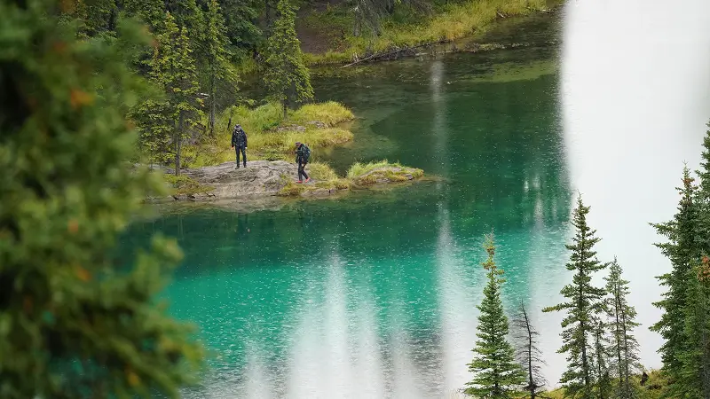 Two hikers standing on a rocky shoreline beside a turquoise mountain lake surrounded by evergreen trees