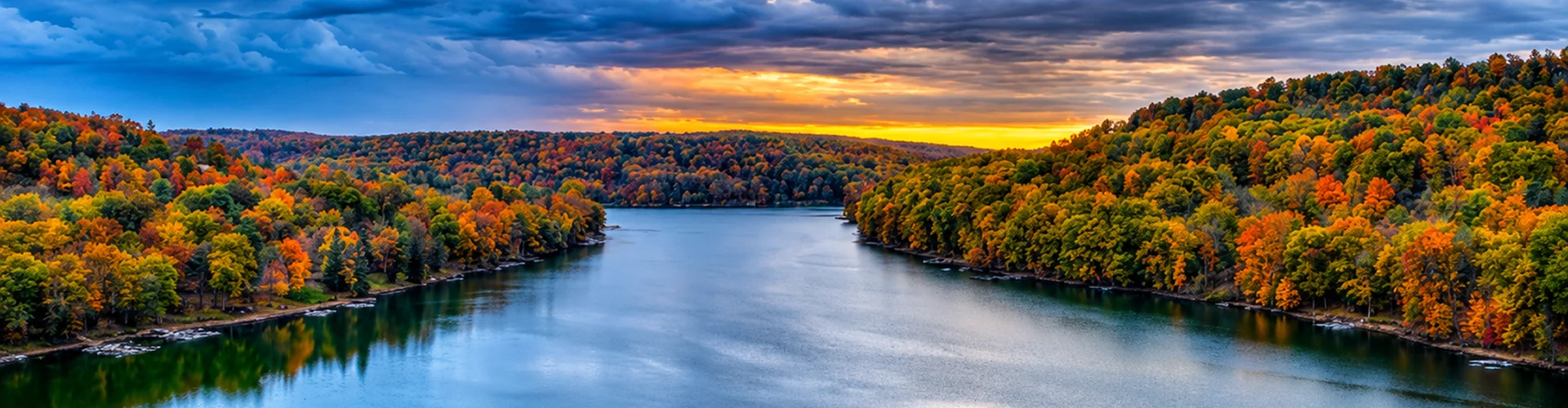 Aerial view of a lake winding through hills covered in vibrant fall foliage, with a colorful sunset on the horizon.