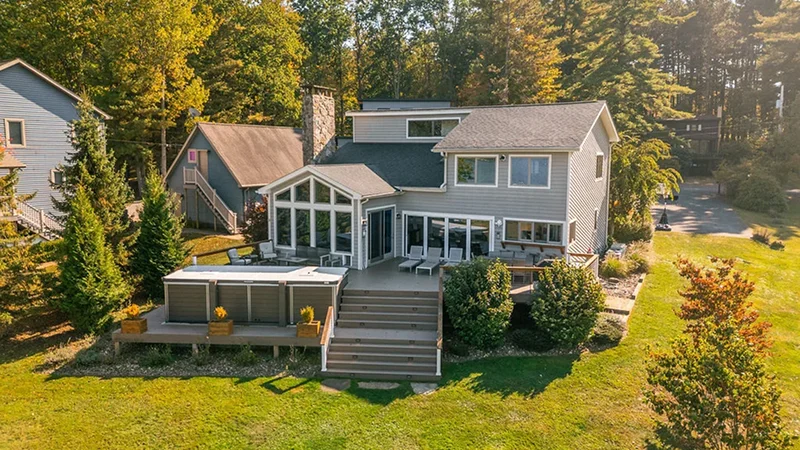 Aerial view of a large vacation home with a deck, hot tub, and lawn surrounded by trees.