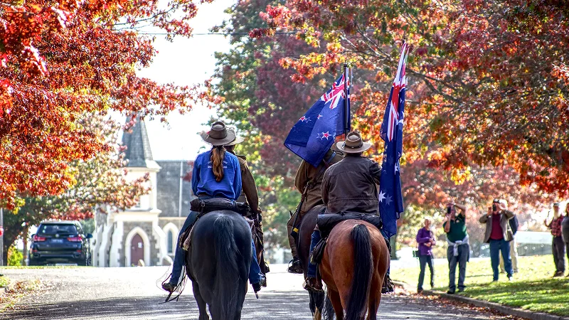Two people on horseback carrying flags ride down a street lined with trees in red and orange fall foliage, with a church in the background.