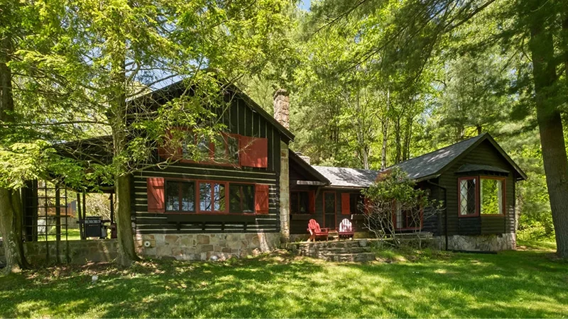 A rustic log cabin with red-shuttered windows and a stone foundation, surrounded by tall green trees.
