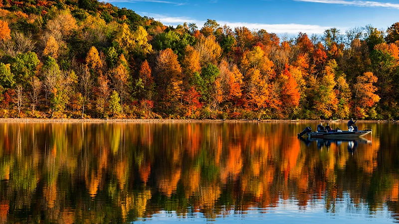 Trees in full autumn color reflecting on a still lake, with a small boat carrying people near the shore.