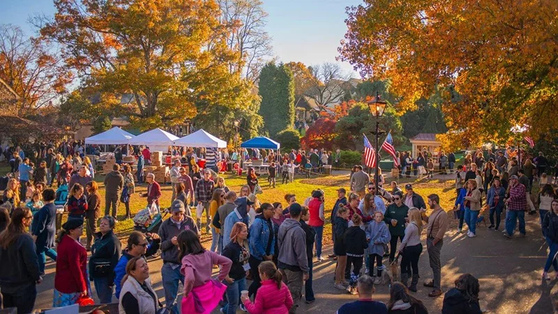A large crowd gathered outdoors under golden fall trees at a festival, with tents and American flags in the background.