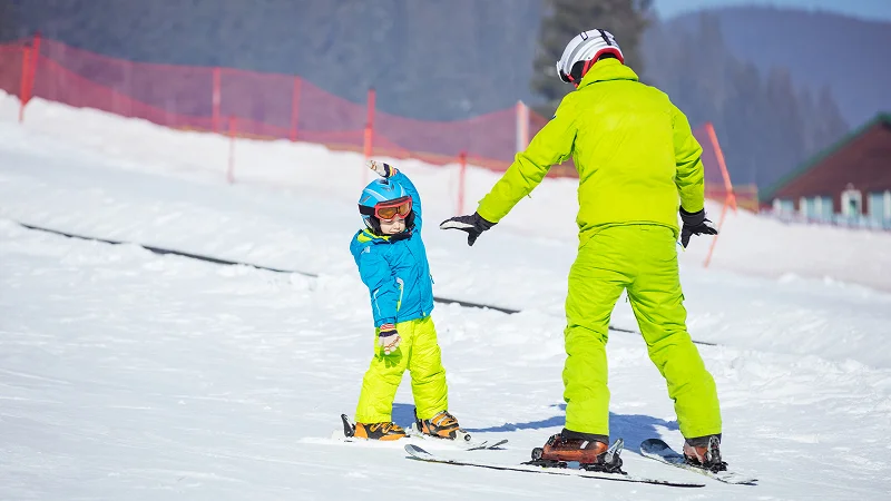 Adult in a yellow ski suit helping a child in a blue jacket learn to ski